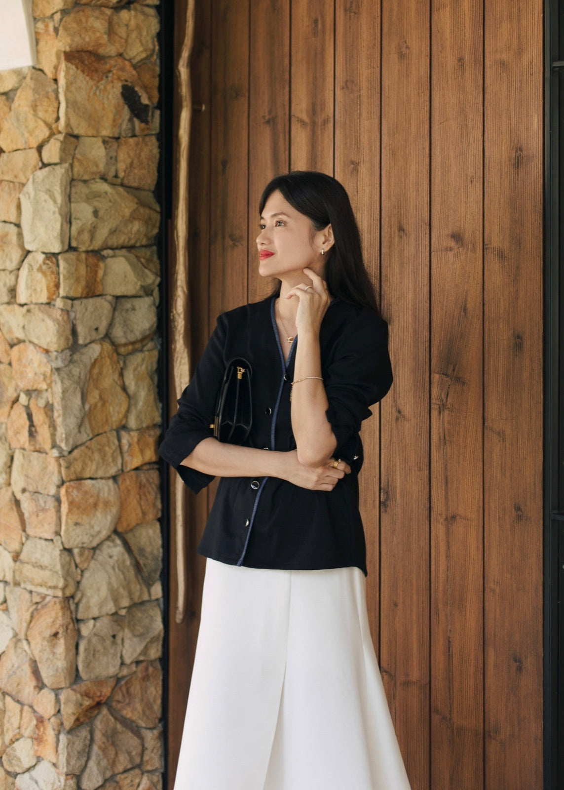 Woman in black blazer and white pants standing against a wooden wall.