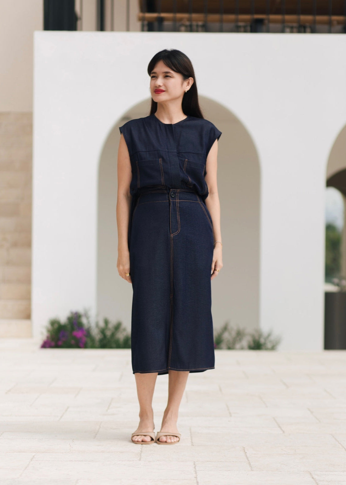 Woman wearing a dark blue sleeveless top and high-waisted pants standing in front of a white wall with arches.