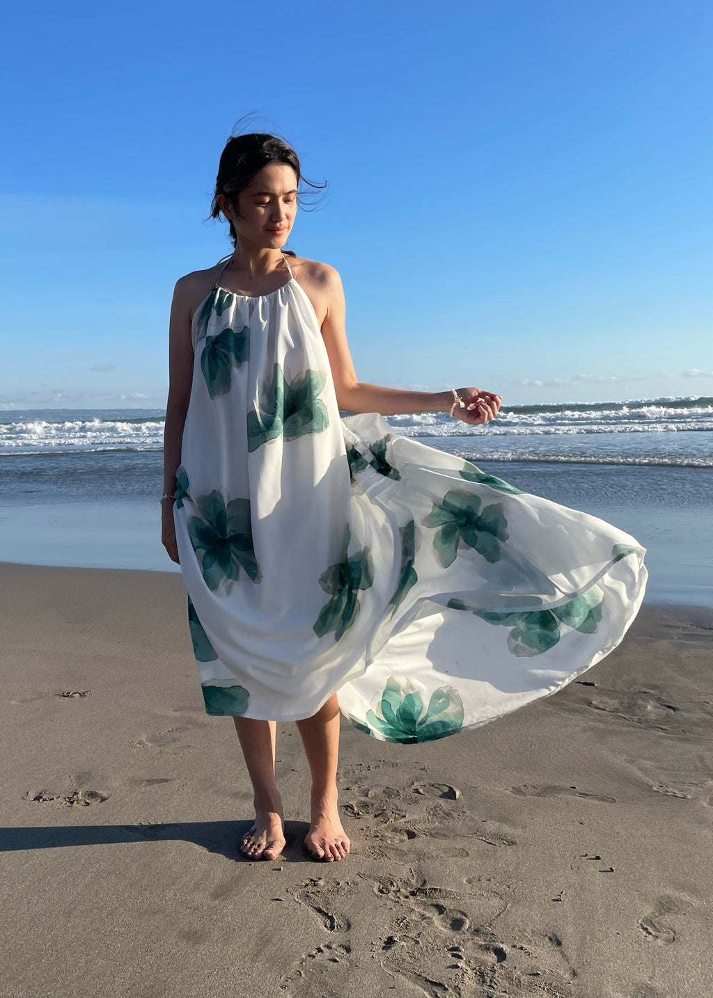 Woman in a white dress with green floral patterns standing on a sandy beach.