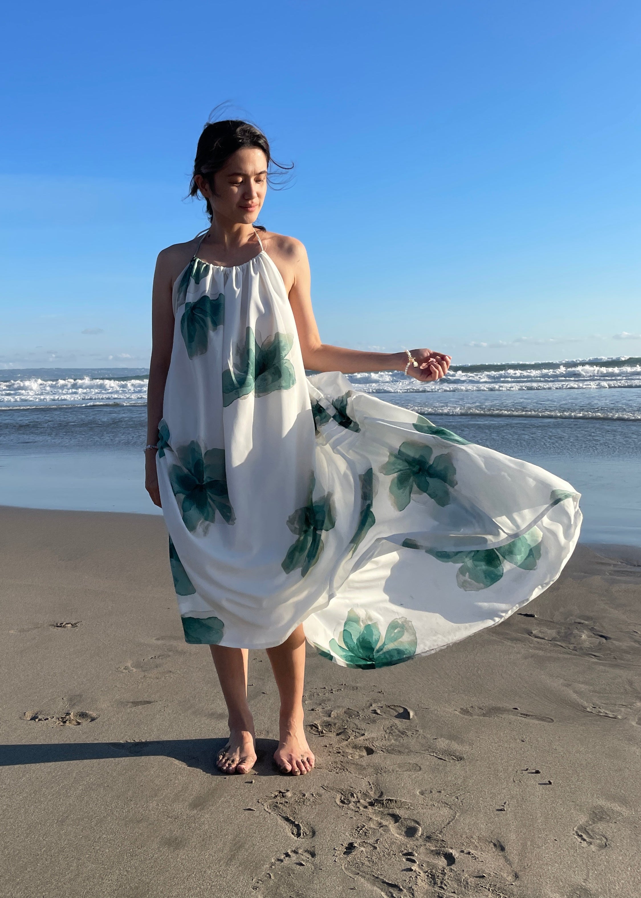 Woman in a white dress with green floral patterns standing on a sandy beach.